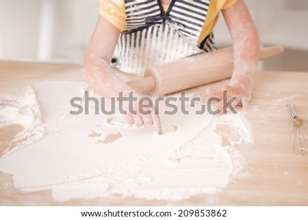 Closeup portrait of cute little girl working with rolling pin in the kitchen wearing apron and drawing sun with flour on kitchen table