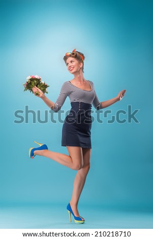 Full length side view portrait of beautiful sexy girl with pretty smile in pinup style wearing navy color dress and sunglasses posing with bouquet of flowers lifting up one leg isolated on blue