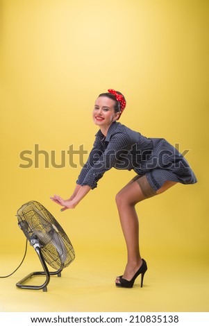 Full length side view portrait of beautiful sexy girl with pretty smile and polka dots dress in pin-up style posing with blowing fan isolated on yellow