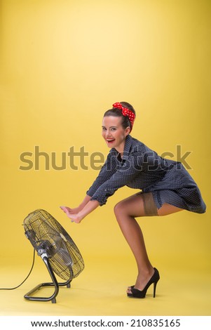 Full length side view portrait of beautiful sexy girl with pretty smile and polka dots dress in pin-up style posing with blowing fan isolated on yellow