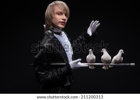 Half-length portrait of fair-haired matchless juggler wearing interesting black costume and white shirt standing aside holding three pouters on the silver stick. Isolated on black background