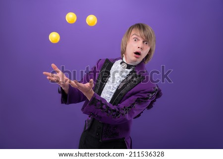 Half-length portrait of funny surprised juggler wearing splendid violet jacket standing aside juggling with the little yellow balls. Isolated on blue background