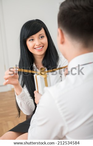 Back view of young businessman giving white gift box to pretty colleague sitting on sofa in office ,woman happy looking at him and opening gift box