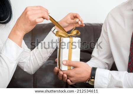 Close-up  view of hands of man giving white gift box, female hands opening gift box in office interior