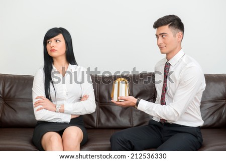 Side view of Business couple in formal clothing sitting on sofa in office, man flirting and giving white gift Box to dissatisfied and unhappy woman that looks aside
