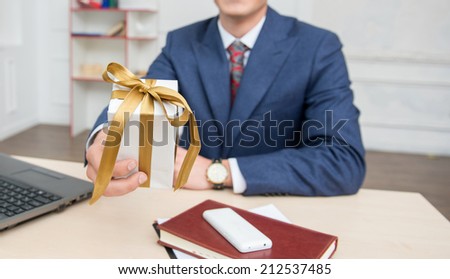 Portrait of young smiling businessman  with a present box in office sitting on the desk working on computer, looking at camera, selective focus on a gift box