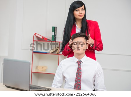 Portrait of young business woman looking at the camera and doing head massage to her colleague with closed eyes sitting at the desk in office with copy space, relationship at work concept