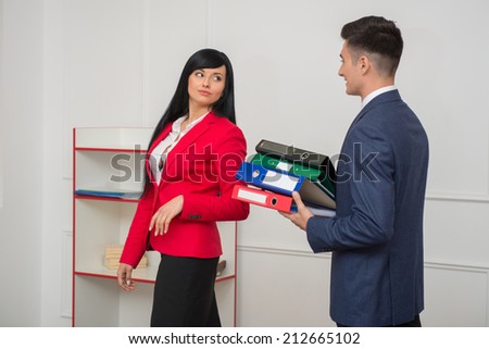 Side view of business man with folders flirting in office with his colleague beautiful lady in red jacket looking at him, with copy space on the wall