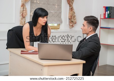 Side view portrait of young business couple flirting near the table at workplace in office, man sitting at the desk and looking at sexy standing colleague, relationship at work concept