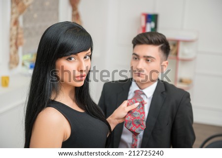 Portrait of young charming businesswoman flirting and pulling her colleague by the tie in office, looking at the camera