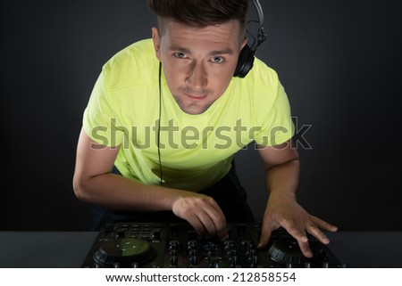 Closeup portrait of confident young DJ with stylish haircut and headphones on head mixing music on mixer looking at camera while standing isolated on dark background