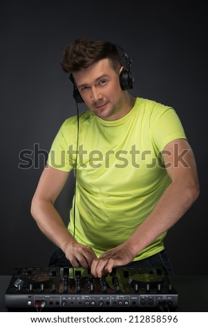 Confident young DJ with stylish haircut and headphones on head mixing music on mixer looking at camera while standing isolated on dark background