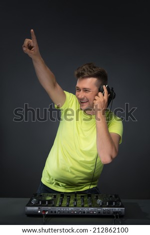 Half-length portrait of happy smiling young DJ with stylish haircut and headphones lifting his finger mixing music on mixer while standing over dark background