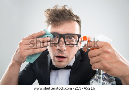 Funny half-length portrait of stylish hipster man in tuxedo and bowtie in spectacles cleaning his glasses with duster, isolated on white