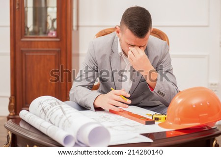 Close-up portrait of young handsome architect engineer working on design plan thinking sitting at the table with safety helmet, spirit level and measuring tape, design bureau background