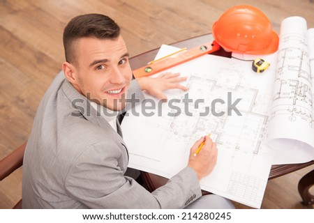 Top view portrait of young smiling handsome architect engineer working on design plan sitting at the table with safety helmet, pencil, spirit level and measuring tape on table in design bureau