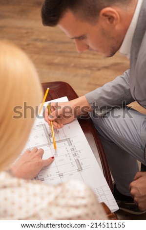 Top view portrait of young handsome architect discussing ground plan with client sitting at the table in design studio, pointing at architectural plan with pencil