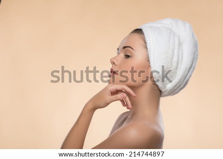 Portrait in profile of beautiful girl in spa with clean and  fresh skin, dreamly looking aside and touching her face with a towel on her head , isolated on beige background with copy place