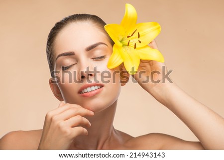 Portrait of beautiful smiling girl with clean and  fresh skin in spa with closed eyes holding yellow  flower near her face and touching her skin isolated on beige background