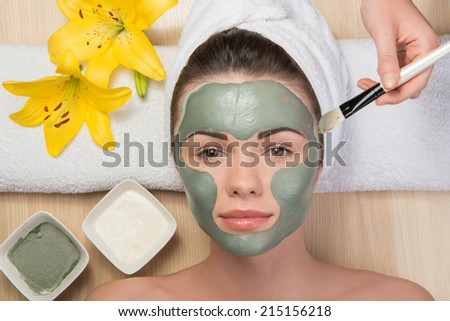 Close-up portrait of beautiful girl looking at the camera with a towel on her head applying facial clay mask and beauty treatments lying on a table in spa near yellow flower and two plates