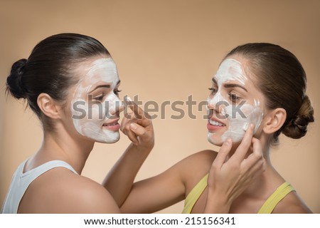 Portrait of two beautiful girls with cream on their  faces looking on each other  and touching their noses with facial cream on their fingers  isolated on beige background with copy place