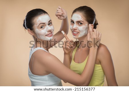 Portrait of two beautiful girls with cream on their  faces looking at the camera  and touching face of each other  with facial cream on their fingers  isolated on beige background