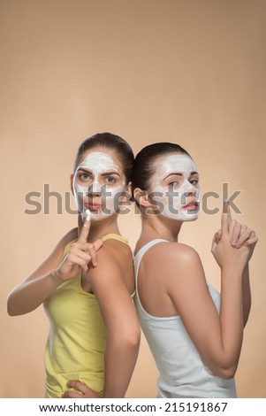 Vertical portrait of two beautiful girls with cream on their faces standing back to back , one girls showing sign attention and other girl showing a gun isolated on beige background with copy place