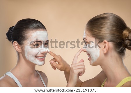 Portrait of two beautiful girls with cream on their  faces looking on each other  and touching their noses and laughing  isolated on beige background with copy placer