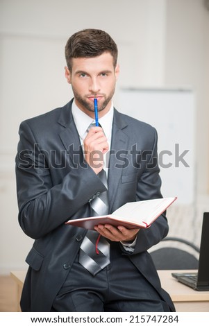 Waist-up portrait of handsome businessman in office interior holding a red notebook in his hands touching his lips with a pen and looking at the camera and thinking, making some decision