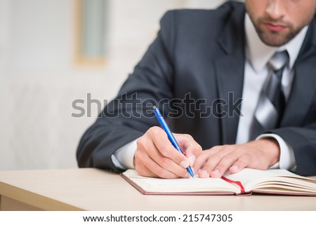 Close-up image of red notebook and hands of businessman sitting at the table in office with a pen attentively writing some notes with copy place