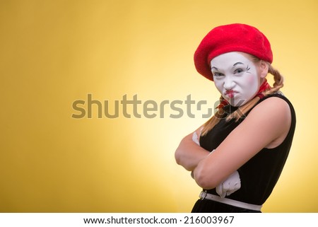 Close-up portrait of playful female mime with white funny face, red hat and red scarf looking at the camera isolated on yellow background with copy place