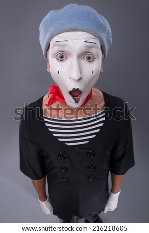 Top-view portrait of very surprised female mime with opened mouth in red head and with white face looking at the camera isolated on grey background