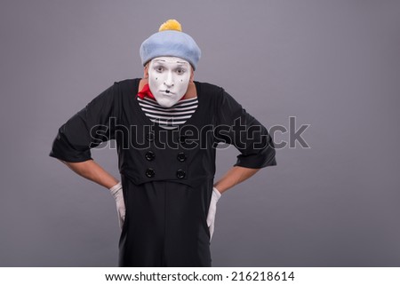Waist-up portrait of funny male mime with grey hat and white face looking at the camera with great surprise and holding his hands on his waist isolated on grey background