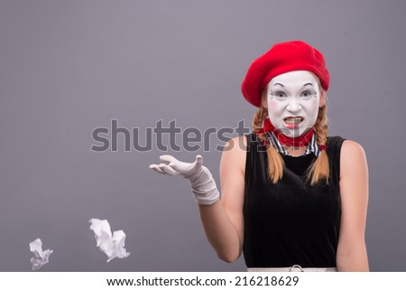 Close-up Portrait of female mime with red hat and white face throwing a crumpled paper and angry looking to the camera isolated on grey background with copy place