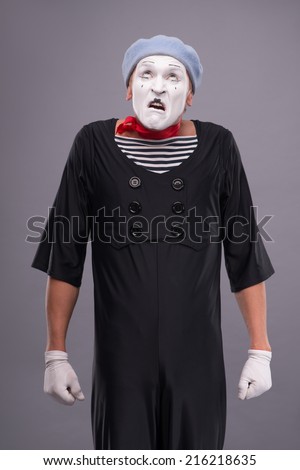 Waist-up portrait of funny male mime with grey hat and white face disappointed looking up and holding his hands omitted down isolated on grey background with copy place