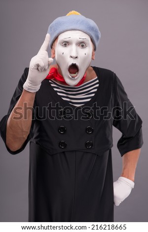 Waist-up portrait of funny male mime with grey hat and white face playfully looking at the camera and showing sign Attention with opened mouth isolated on grey background with copy place