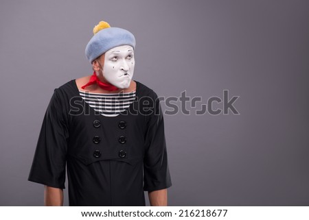 Waist-up portrait of funny male mime with grey hat and white face disappointed looking up and holding his hands omitted down isolated on grey background with copy place