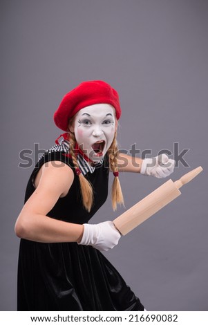 Waist-up portrait of female mime with red hat and white face screaming and holding a rolling pin isolated on grey background with copy place
