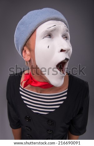 Top-view close-up portrait of funny and surprised male mime with opened mouth in red head and with white face looking aside isolated on grey background