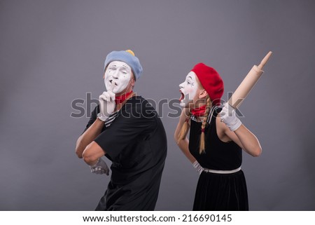Waist-up portrait of mime couple with white faces, female mime wanting to beat male mime with rolling pin and male showing sign Silence isolated on grey background with copy place