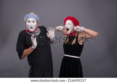 Waist-up portrait of funny mime couple with white faces, female mime closing her ears with her fingers and shouting on male mime carefully waving his hand isolated on grey background