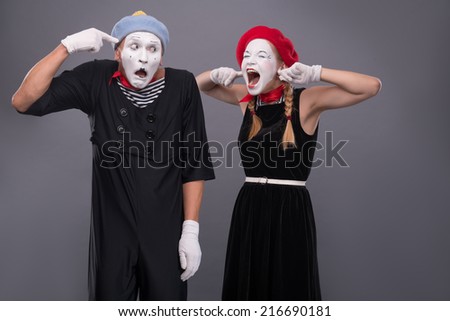 Waist-up portrait of mime couple with white faces, female mime closing her ears with her fingers and shouting on male mime showing that she is crazy isolated on grey background