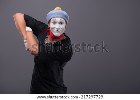 Waist-up portrait of funny male mime with grey hat and white face happy looking at the camera with great surprise and holding his hands on the chest isolated on grey background with copy place