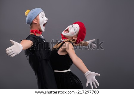 Waist-up portrait of mime couple wanting to hug each other, female mime looking at the camera, male mime laughing and looking at his girlfriend isolated on grey background with copy place