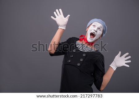 Waist-up portrait of funny male mime with grey hat and white face happy looking up with great surprise and breeding his hands isolated on grey background with copy place