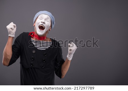 Waist-up portrait of funny male mime with grey hat and white face showing sign YES with both hands and closing his eyes and smiling isolated on grey background with copy place