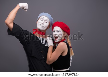 Waist-up portrait of funny  hugging mime couple, male mime looking at his girlfriend and showing his strength, female mime looking at the camera, isolated on grey background with copy place