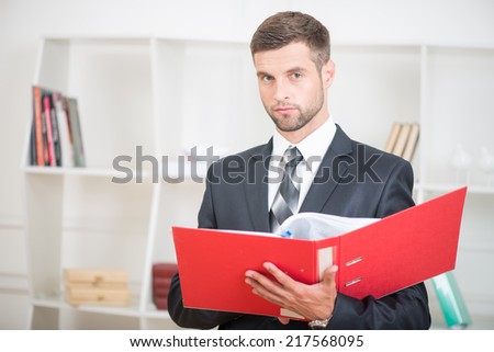 Close-up portrait of handsome confident businessman standing in office with a red folder in his hands and looking at the camera with serious face