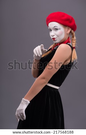 Waist-up portrait of young mime girl with white face, red hat and red scarf sexy looking at the camera and showing at you isolated on grey background with copy place
