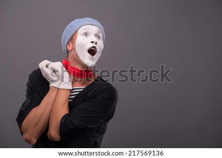 Waist-up Portrait of young male mime holding hands near his face like a girl and playfully looking aside and singing isolated on grey background with copy place